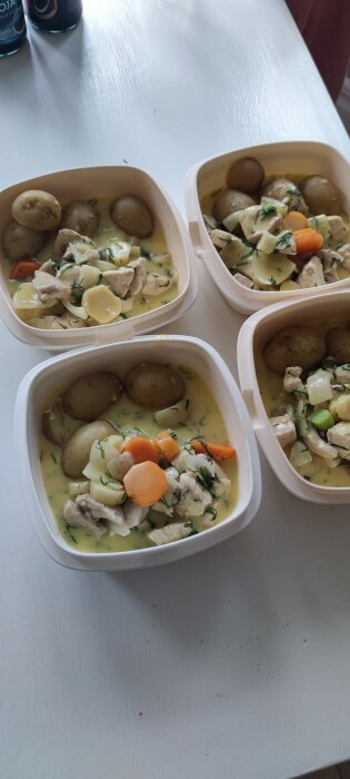 Fyra matlådor med dillkyckling, potatis och morötter i sås, på ett vitt bord.
