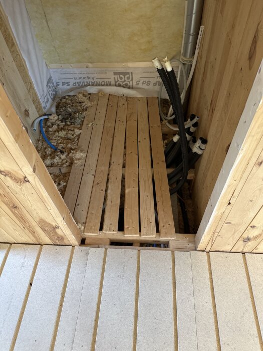 Trägolv med råspont och synliga rördragningar i bjälklag under renovering, samt isoleringsmaterial på golvet.