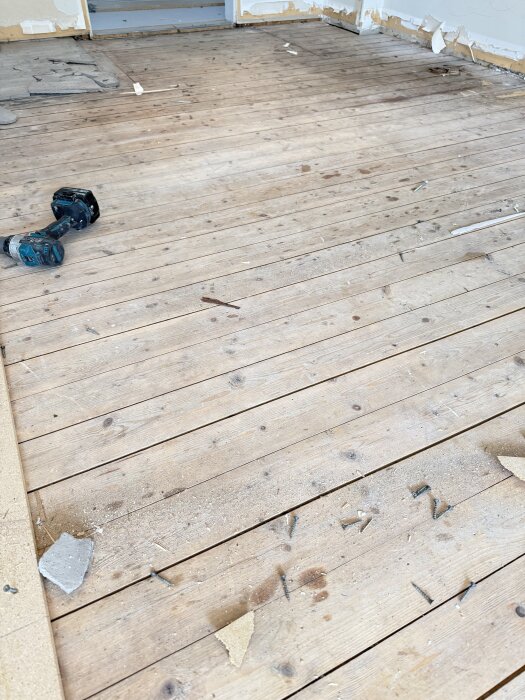 Gammalt furugolv med slitna plankor, skruvar och byggdamm synliga. En borr ligger på golvet i ett rum under renovering.
