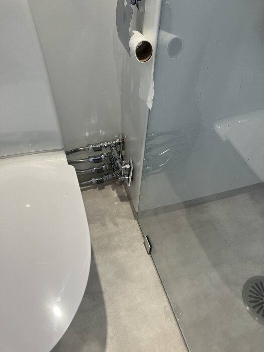 Rörgenomföring i badrumsvägg vid dusch med utanpåliggande kopplingar och grå våtrumsmatta från Tarkett; toalett och dusch synliga i bakgrunden.