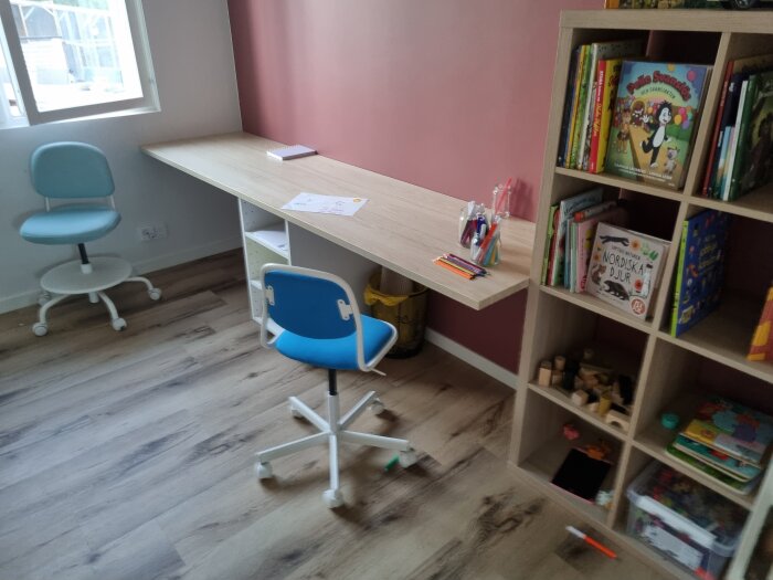 Pysselrum med rosa vägg och träbänk, två olika stolar, hylla med barnböcker och leksaker, skrivbordsmaterial spridda över bänkskivan.