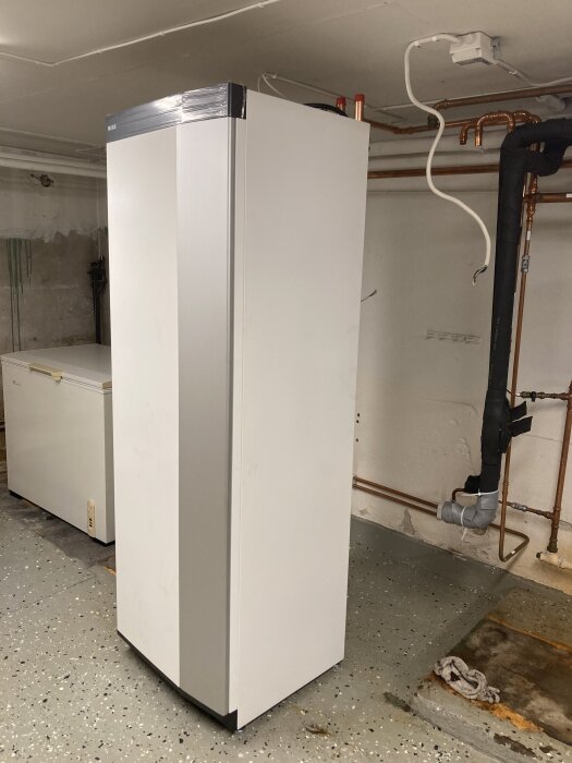 Ny vit vattenpump installerad på betonggolv i källare, omgiven av kopparrör och elkablar.