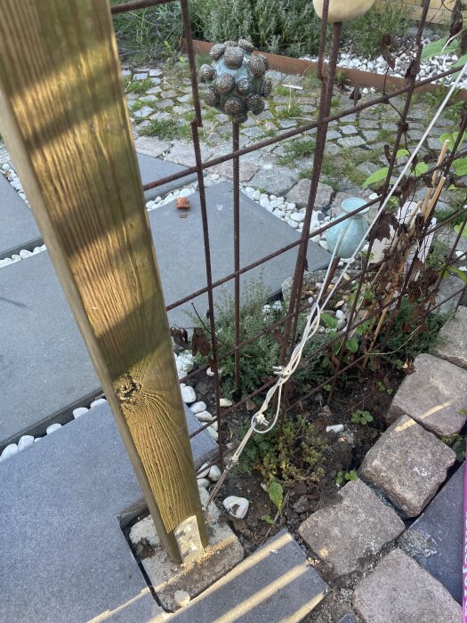 Trädgårdsinstallation med vantskruv och vajer vid trästolpe, omgiven av växtlighet och stenläggning, del av en grindkonstruktion.