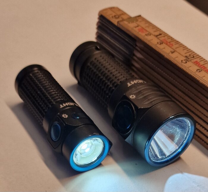 Två ficklampor, Olight Baton 4 och Olight Baton Turbo, bredvid en hopfällbar tumstock på vit bakgrund.