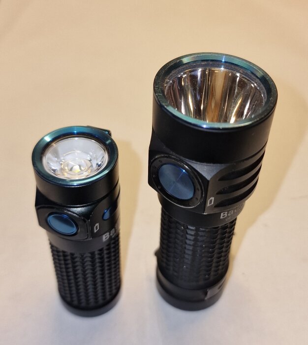Två ficklampor stående på ett bord; den mindre Olight Baton 4 och den större Olight Baton Turbo, båda med texturerade kroppar och stora knappar.