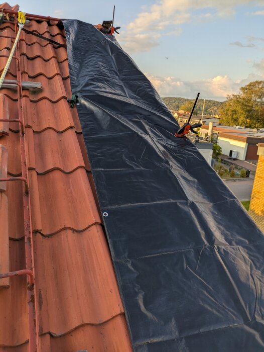 Svart presenning täcker en del av taket med tegelpannor, fastsatt med klämmor. I förgrunden syns en säkerhetsselens krok.