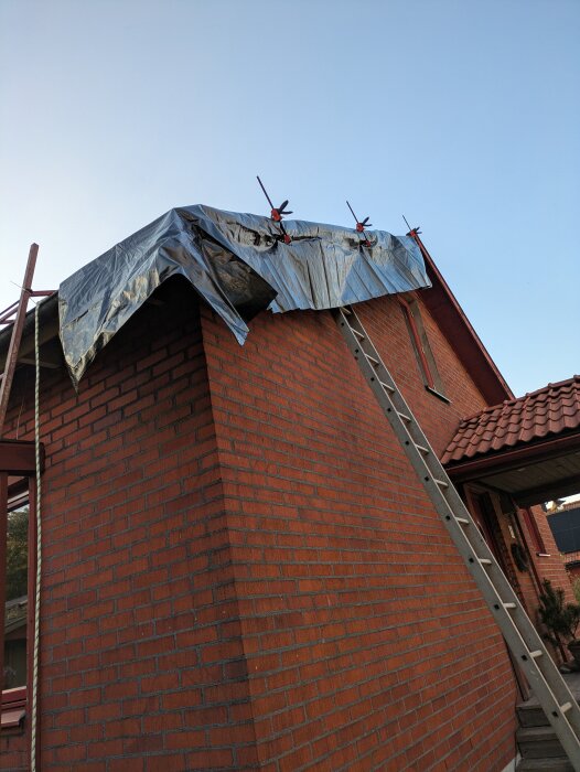 Röd tegelvägg med presenning fastsatt med klämmor på taket; stege lutad mot väggen.