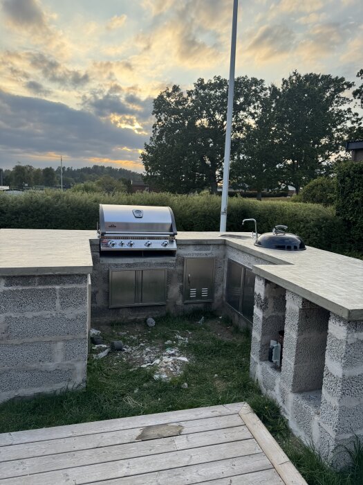 Utomhuskök med monterad grill och diskho på cementbänk, omgiven av gräsmatta och träd under molnig himmel.