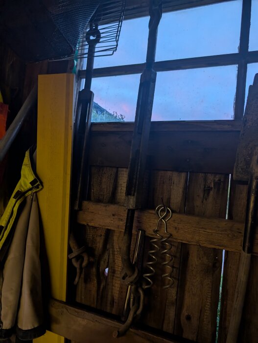 Vantskruv hänger vid ett fönster i ett verktygsskjul, solnedgång syns utanför.
