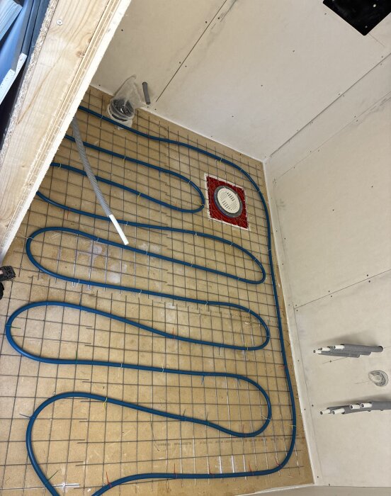 Plastslang lagd över ett metallnät med brunn i ett badrum under renovering, synligt spårade skivor och plåtar för golvvärmeinstallation.