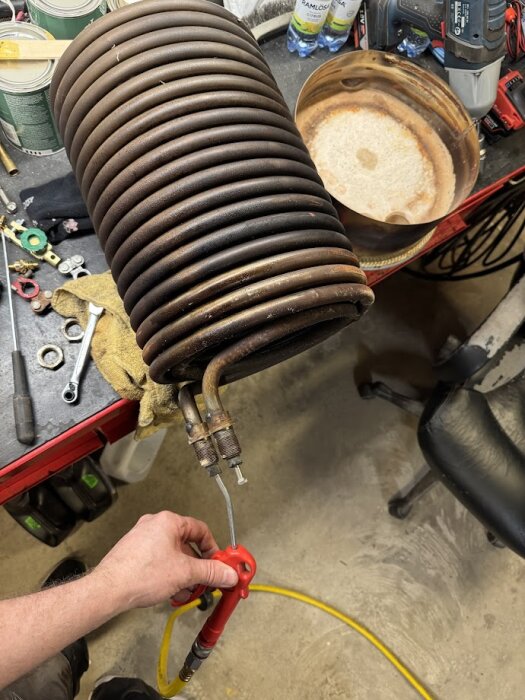 En person håller en tryckluftspistol vid en spiralformad värmeslinga på en arbetsbänk med verktyg.