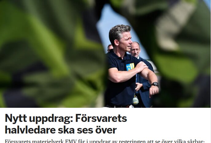 En man i blå t-shirt talar utomhus med flera personer i bakgrunden, delvis dolda av gröna löv.