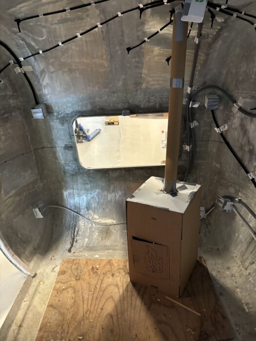 Kartongmodell av bastuaggregat placerad på trägolv i en metallvagn. Modellen har en ritad eldsymbol och ett rör uppåt mot taket.