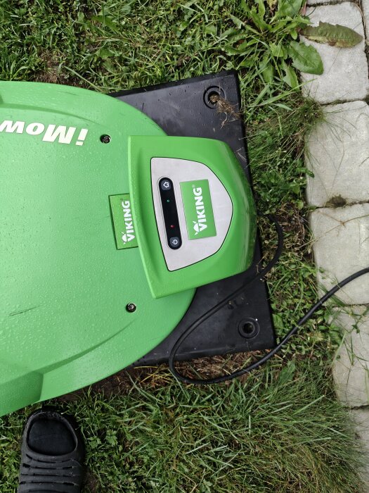 Grön robotgräsklippare från Viking på laddningsplatta i en trädgård, omgiven av gräs och stenar.