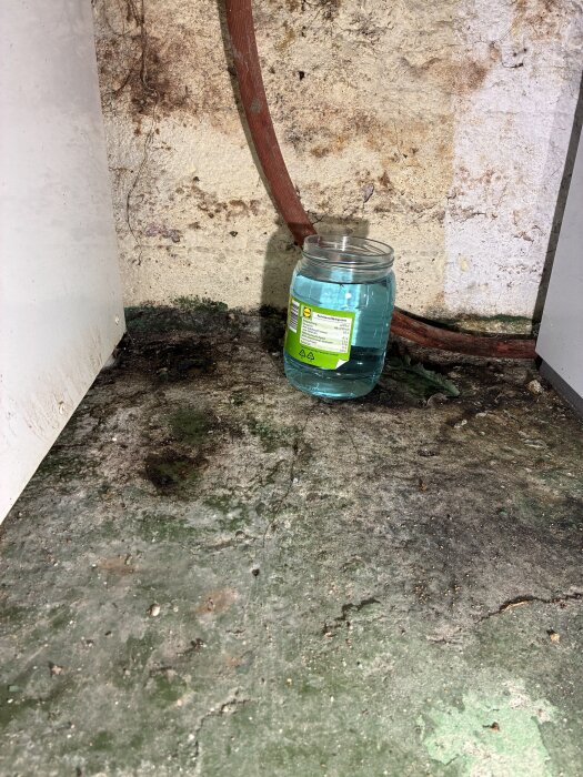 Glasburk fylld med blå vätska placerad under ett rör, stående på ett slitet och fuktigt golv, indikerar läckage från bergvärmepumpsystem.