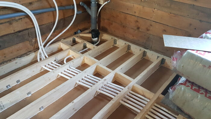 Rör och träreglar under golv i badrumsrenovering, med slangar som leder ner till källarens fördelningsrör.