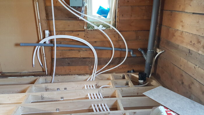 Badrumsrenovering med oskarvade slangar som går ner till fördelningsrör. Träribbor på golvet och väggar, samt flera synliga rör och fönster.