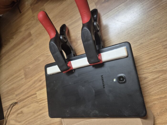 Samsung-surfplatta med en trälist fastspänd för stabilitet, med två klämmor på ett trägolv.