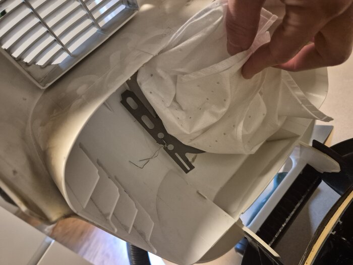 En hand fäster dammsugarpåsen med ståltråd i dammsugaren, där en plastbygel saknas, för att anpassa olika påsmodeller.