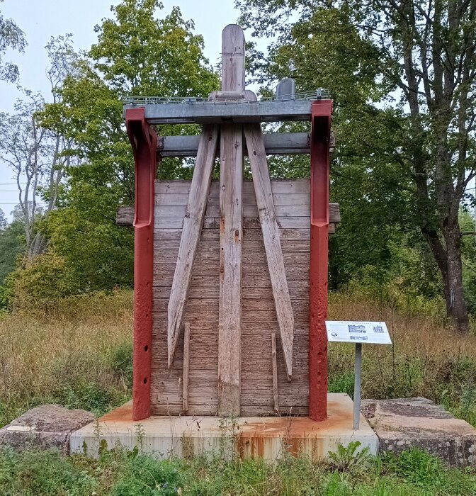 Gamla dammluckan vid Hyndevadsdammen med trästruktur och röda metallstöd, omgiven av träd och gräs.