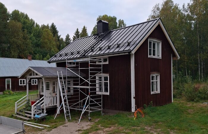 Rött timmerhus med nytt svart plåttak, omgiven av byggnadsställning och verktyg, grönska och ett äldre mindre tak på husets framsida.