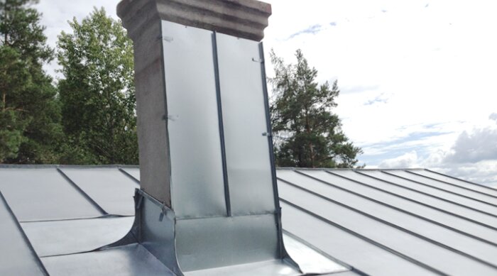 Skorstensrör med metallskydd på ett plåttak, omgiven av grönskande träd och en klar himmel.