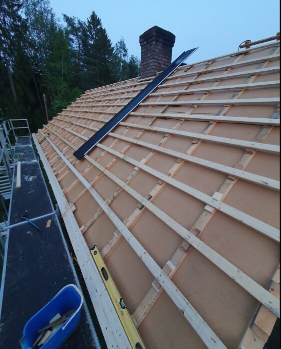 Tak under renovering med ny strö- och bärläkt monterad, synliga byggställningar runt huset. Stege och vattenpass ligger på taket.