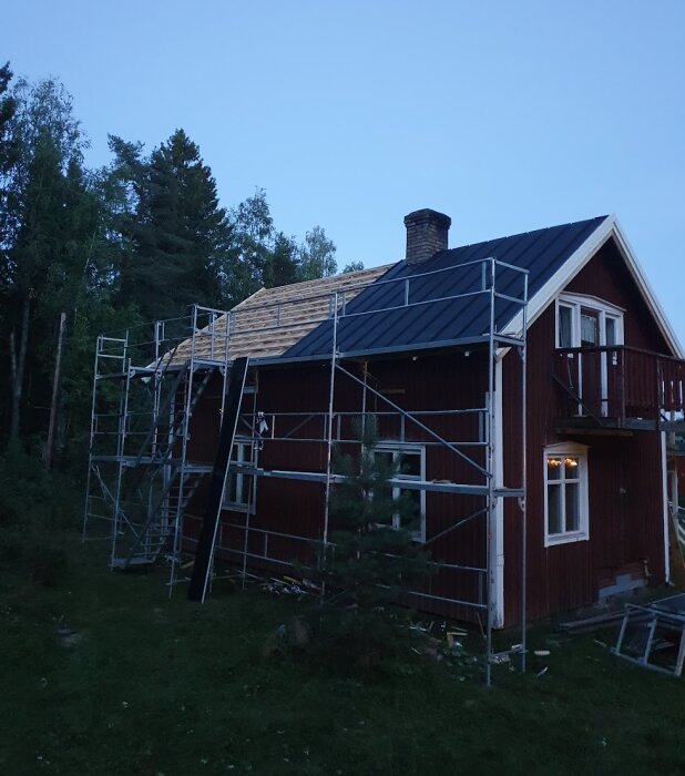 Rött timmerhus med monterad byggställning under takrenovering, delvis nytt tak av träbalkar och metallplattor, skog i bakgrunden.