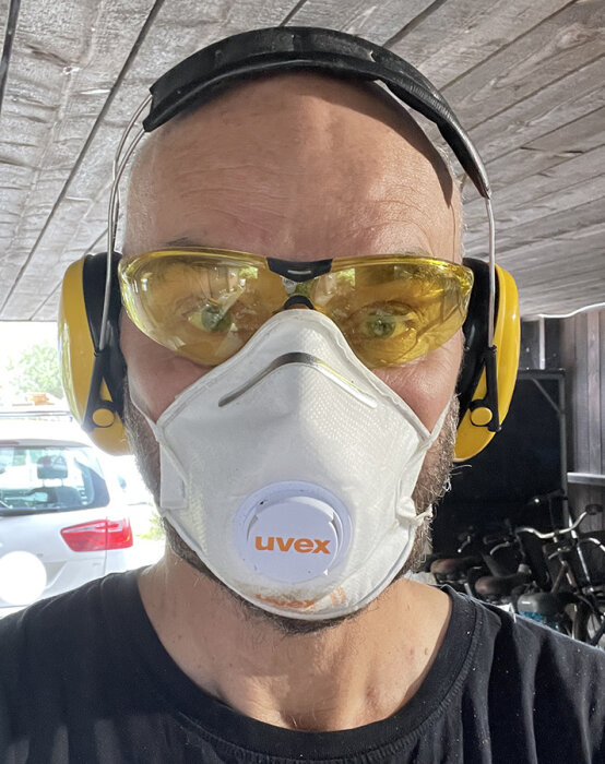 Person med hörselskydd, skyddsglasögon och andningsmask arbetar med träprojekt. Bakgrund med bil och trästruktur.