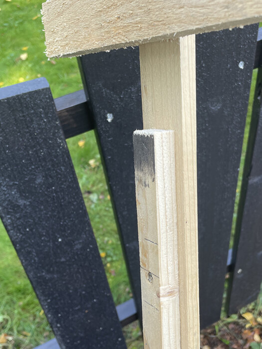 Mall för att byta staketbrädor, med snedsågad bräda och markeringar för skruvplatser, mot en bakgrund av svart staket.