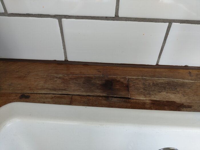 Sliten träbänkskiva vid diskho med mörk ytskada, omgiven av vitt kakel och silikonfog.