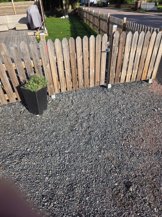 Trädgårdsgrind av trä med småsten på marken, omgiven av mulched yta och gräs med en uteplats i bakgrunden.