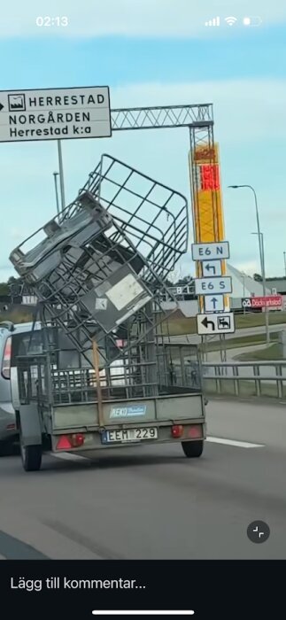 Släpvagn på motorväg med högt staplade metallställningar fastsurrade, vägvisare för Herrestad i bakgrunden.