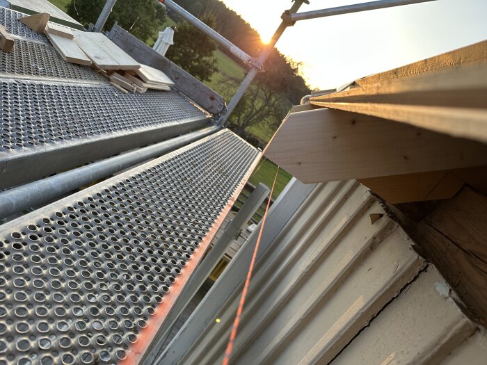 Byggnadsställning med plattform och träplankor under montering för takfotsförlängning vid solnedgång, med laserlinje längs taket.