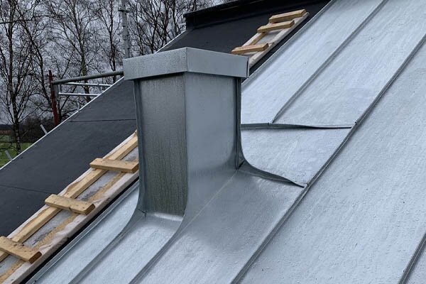 Plåttak med en skorstenshuv och träreglar på en lutande takyta, omgivna av träd i bakgrunden.