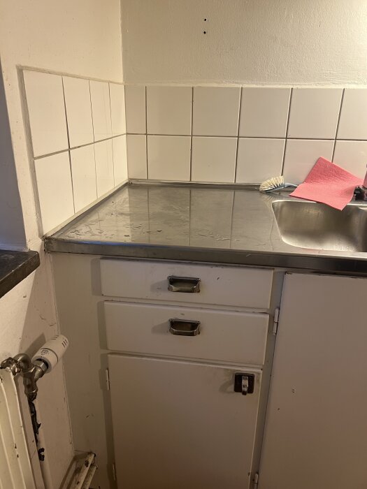 Kökshörna med diskbänk, kaklat stänkskydd och två borrhål i väggen ovanför. Vänster sida visar elementrör.