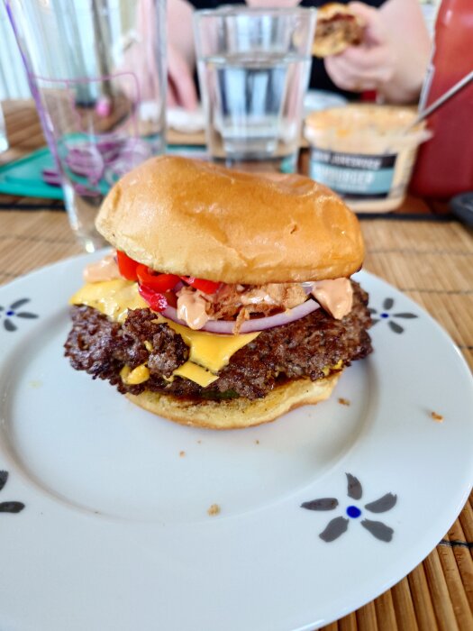 Dubbel hamburgare på tallrik med ost, picklad chili, saltgurka, rödlök och dressing. Bakgrund med glas och dressingburk.
