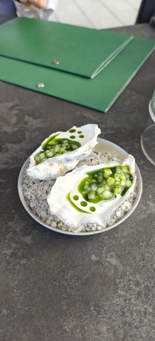 Två ostronskal fyllda med grön sås och tärnade ingredienser på en tallrik med stenar, placerad på ett bord.