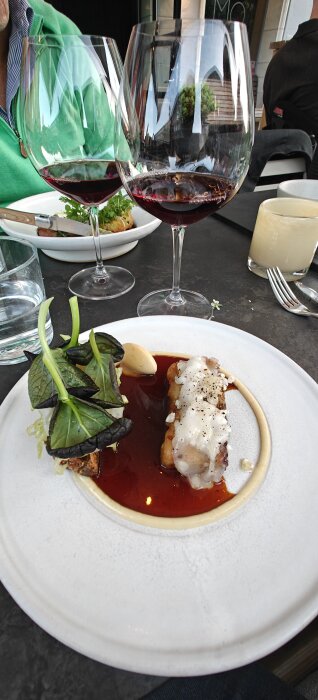 Två glas rödvin och en tallrik med en elegant maträtt dekorerad med gröna blad på restaurang, del av en kvällsupplevelse.