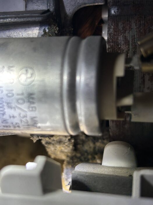 Närbild av en metallisk och plastkomponent i en maskin, med text och viss smuts synlig, förmodligen en del av ett rörsystem eller motor.