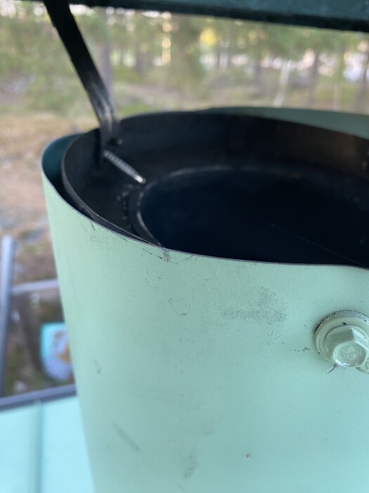 Närbild av en metallisk komponent, troligen en del av en ventilationsrörstruktur, fotograferad från taket. Bakgrund med suddiga träd.