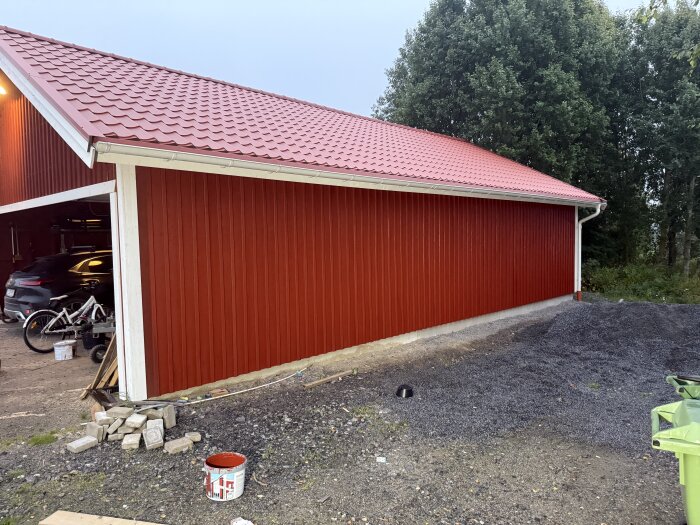 Röd nymålad vägg på en byggnad med vitt undertak och rött tak, omgiven av grusmark; färgburk synlig på marken.