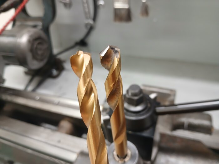 Två guldiga borr, varav ett ser vant slipat ut, står i en metallchuck på en svarv, med suddig bakgrund av verktygsmaskiner.