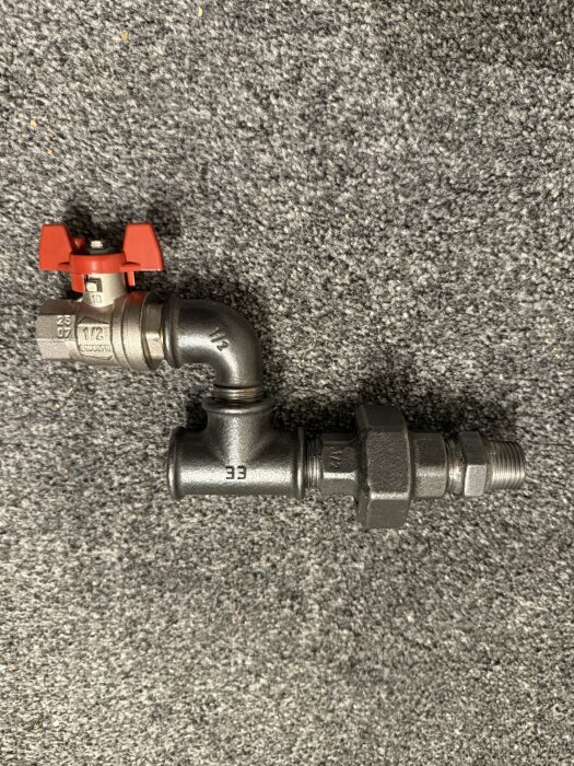 Rörkoppling med metallventil och röd vred placerad på grått underlag, avsedd som ersättning för ett skarvrör.