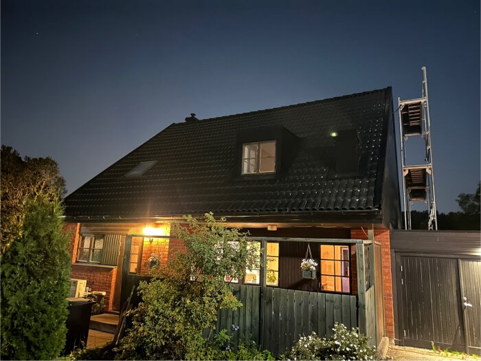 Mörkt foto av ett hus med belysning, där takrännans lutning och pannornas placering syns på taket, pallställning på höger sida.