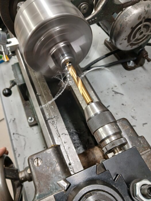 Svarv med borrchuck och borr, där borren bearbetar metall och avger spån, i en verkstadsmiljö.