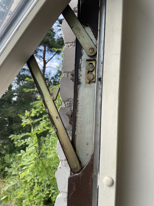 Närbild av ett öppet fönster som visar den mekaniska delen av gångjärnet, omgivet av tegel och en träkarm.