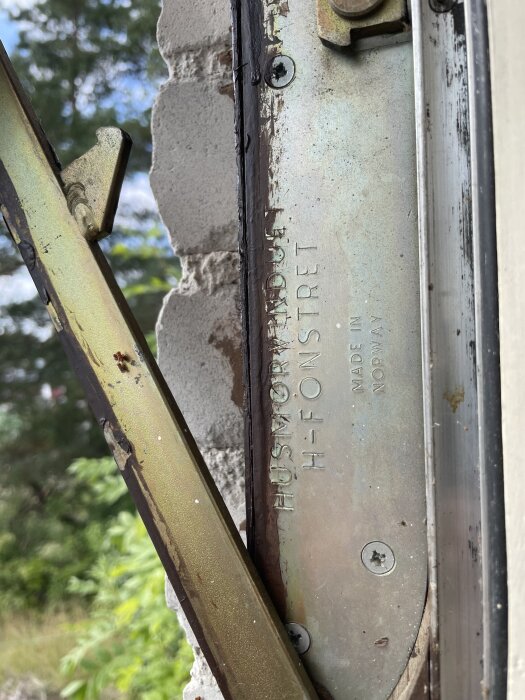 Närbild på beslagen av ett fönster med texten "HUSMODERFÖNSTER" och skruvar, fönstret är delvis omgivet av tegel och har natur i bakgrunden.