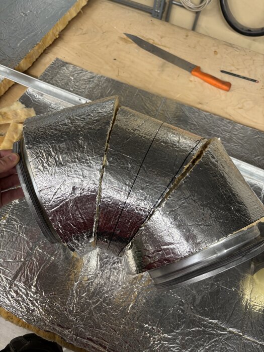 Silverisolerad 90 graders böj av lamell med tre sektioner, placerad på en arbetsbänk med verktyg i bakgrunden.