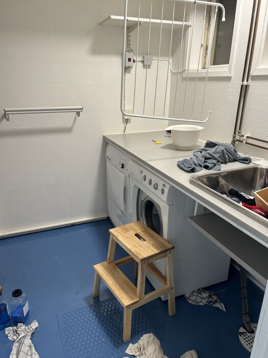 Tvättstuga med torktumlare, diskho och träpall. Blått golv med vattenproblematik och handdukar på golvet för att torka upp.